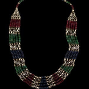 May include: A multi-strand necklace featuring red, green, and blue beads. The necklace has silver-toned metal accents and a chain clasp. The beads are arranged in vertical rows, creating a layered effect. The background is black.