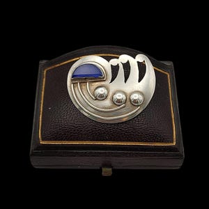 Puede incluir: Broche Art Déco plateado con un acento de piedra azul y tres pequeñas esferas plateadas. El broche se muestra en un joyero marrón oscuro con ribete dorado. El diseño es geométrico y estilizado, con una forma curva.