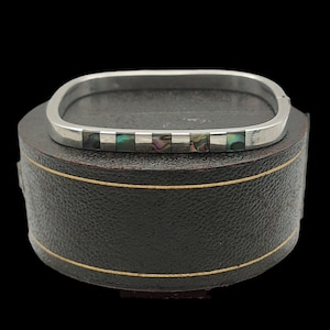 May include: A silver bangle bracelet with a square shape and a row of iridescent abalone shell inlay.