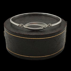 Peut inclure: Un bracelet jonc argenté avec une pièce maîtresse ovale. La pièce maîtresse a un petit accent rond. Le bracelet est présenté sur une boîte ronde marron foncé avec une bordure dorée. Le fond est noir.