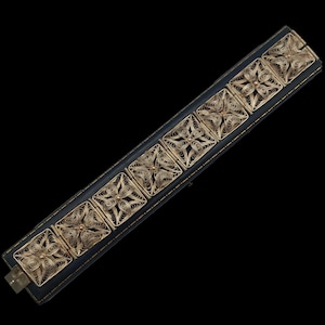 May include: A silver filigree bracelet with square links, each featuring an intricate floral design. The bracelet is displayed on a dark blue rectangular box with a black background. The bracelet is a vintage piece.