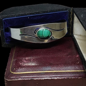 May include: A silver bangle bracelet with a green malachite stone set in the centre. The bracelet is in a velvet-lined box.