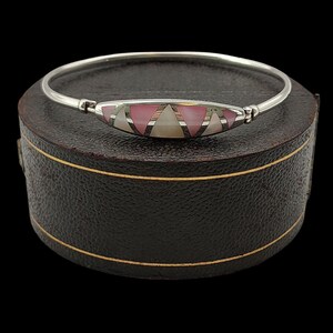 Könnte beinhalten: Ein silbernes Armreif-Armband mit einem zentralen dekorativen Element. Das Element weist rosa, weiße und braune geometrische Formen auf. Das Armband wird auf einer dunkelbraunen, lederartigen Schachtel mit goldenem Rand vor schwarzem Hintergrund präsentiert.