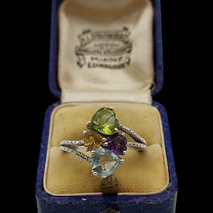 May include: A silver ring featuring heart-shaped gemstones in green, yellow, purple, and blue. The ring is displayed in a vintage blue jewellery box with a cream interior.