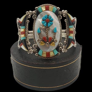 May include: Silver cuff bracelet with an oval mother-of-pearl center featuring a turquoise flower and red accents. The bracelet has black, red, and turquoise geometric designs on the sides. The bracelet is displayed on a black box.
