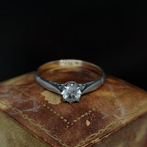 Op de afbeelding: Een zilveren ring met een ronde, heldere edelsteen, gezet in een vier-klauwzetting. De ringband is dun en heeft een gouden binnenkant. De ring wordt gepresenteerd op een bruin leren oppervlak.