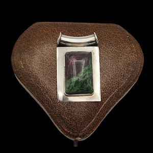 Puede incluir: Un colgante de plata con una piedra rectangular multicolor con tonos morados y verdes. El colgante está engastado en un marco plateado y tiene una barra curva en la parte superior. El colgante descansa sobre una superficie marrón similar al cuero.