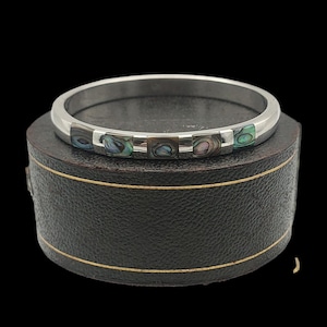 May include: A silver bangle bracelet featuring rectangular abalone shell inlays. The bracelet is presented on a black jewellery box with gold trim. The abalone inlays display iridescent colours of blue, green, and brown.