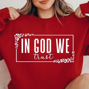 May include: Red sweatshirt with white text that says "In God We Trust" surrounded by a white leopard print design.