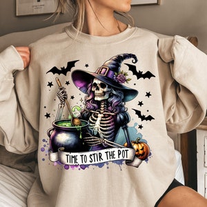 May include: A beige sweatshirt with a graphic print of a skeleton witch stirring a cauldron. The witch is wearing a purple and black hat and has purple hair. The text on the sweatshirt reads "Time to Stir the Pot".