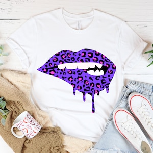 Puede incluir: Una camiseta blanca con un diseño de labios de estampado de leopardo morado y rosa. Los labios gotean con pintura morada.