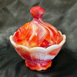 May include: A vintage candy dish with a lid, in red, orange, and white. The glass dish has a scalloped edge and a textured, swirled pattern on the lid. The base is a matching colour and shape.