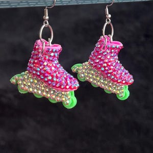 May include: A pair of roller skate earrings. The earrings are shaped like pink roller skates with iridescent rhinestones covering the boot. The wheels are green, and the earrings hang from silver-colored hooks. The background is black.