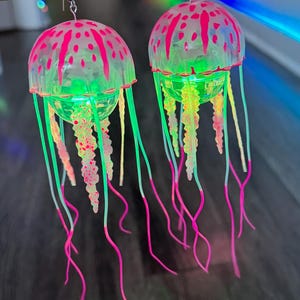 May include: A pair of jellyfish earrings. Each earring features a clear, dome-shaped top with pink accents and a green base. Long, flowing tendrils in pink, green, and yellow extend downward. The earrings are suspended from silver hooks.