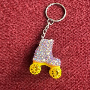 May include: A roller skate keychain with a silver key ring and chain. The roller skate is yellow with yellow wheels and is covered in iridescent rhinestones. The keychain is on a red background.