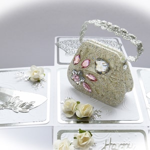 May include: A white and silver layered card with a glittery, beige handbag with a silver chain handle and pink and clear rhinestones. The card also features a white shoe, white flowers, and a silver background.