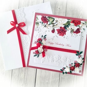 May include: A white card with a red velvet ribbon and lace trim. The card is open to reveal a white card with a red floral design and the text "Happy Birthday Mum".