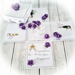 May include: A white explosion box with a silver and white design. The box is decorated with purple flowers and has a message that says "Congratulations on your Engagement".