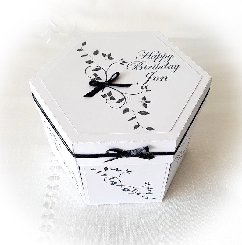 May include: White hexagonal gift box with a black ribbon and a black and white floral design. The box has the text "Happy Birthday Jon" on the top.