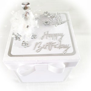 May include: White gift box with a silver ribbon and a silver butterfly decoration. The lid of the box has a silver glitter background and the words "Happy Birthday" in silver lettering.