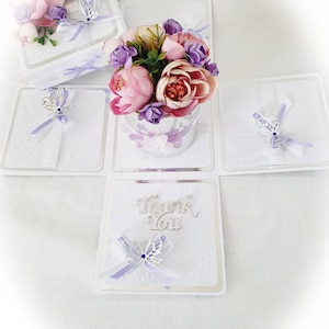 May include: A white card box with a pop-up flower arrangement. The flowers are pink and purple. The box has a silver butterfly and the words "Thank You" on the front.