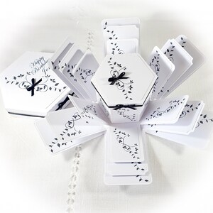May include: White hexagonal explosion box with black ribbon and leaves design. The box is open and the lid has the text "Happy Birthday, Jo."