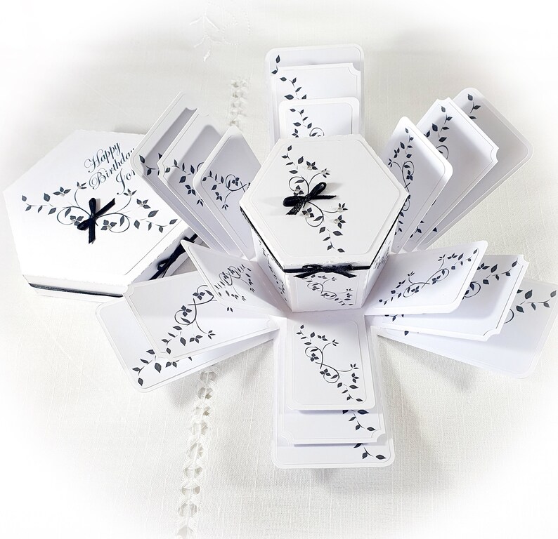 May include: White hexagonal explosion box with black ribbon and leaves design. The box is open and the lid has the text "Happy Birthday, Jo."