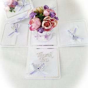 May include: A white square gift box that unfolds into four panels, featuring a central bouquet of pink and purple artificial flowers. Each panel is decorated with a silver butterfly cutout, a light purple ribbon, and the text Thank You in silver.