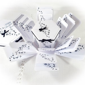 May include: White and black explosion box with a hexagonal center and a black ribbon. The box is decorated with a black and white floral design and the text "Happy Birthday Jon".