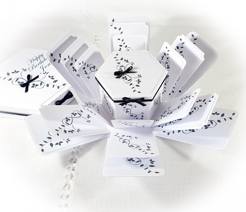 May include: White and black explosion box with a hexagonal center and a black ribbon. The box is decorated with a black and white floral design and the text "Happy Birthday Jon".