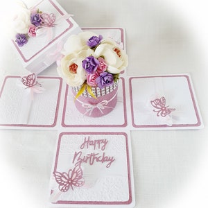 May include: A white and pink explosion box with a pink glitter flower pot and white flowers. The box has a pink border and the words "Happy Birthday" on the front.