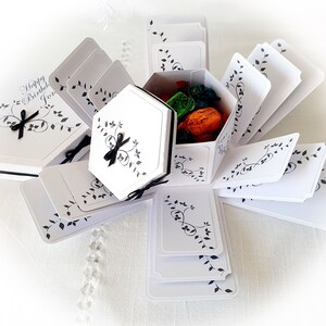 May include: White explosion box with black floral designs and a black ribbon. The box is open and has a small white box inside with a black ribbon. The small box contains colorful candies.