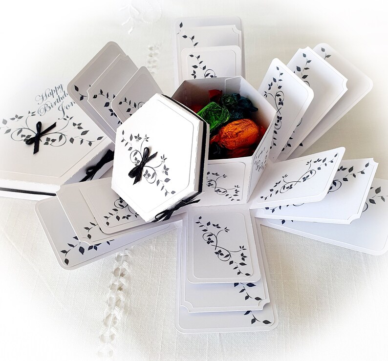 May include: White explosion box with black floral designs and a black ribbon. The box is open and has a small white box inside with a black ribbon. The small box contains colorful candies.