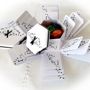 May include: White explosion box with black floral design and the text "Happy Birthday Jon". The box is open and filled with small gifts.