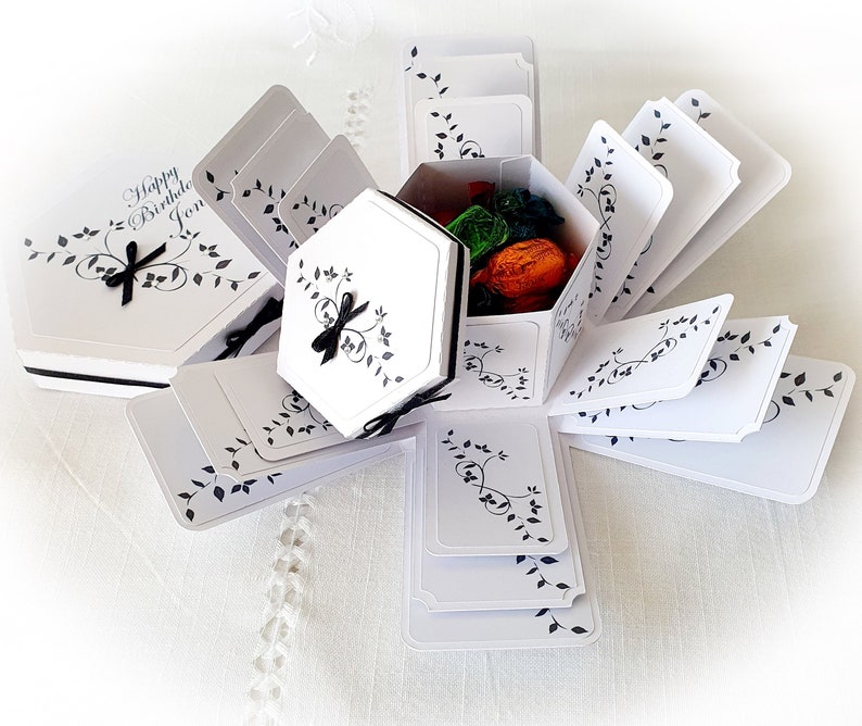 May include: White explosion box with black floral design and the text "Happy Birthday Jon". The box is open and filled with small gifts.