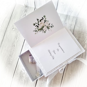 May include: White wedding card with a white floral design on the front and the text "Congratulations on your Wedding Day" on the inside. The card is open to reveal a small white box with a white ribbon and a white flower.