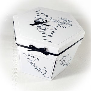 May include: White hexagonal gift box with a black ribbon and bow. The box has a floral design and the text "Happy Birthday Jon" on the top.