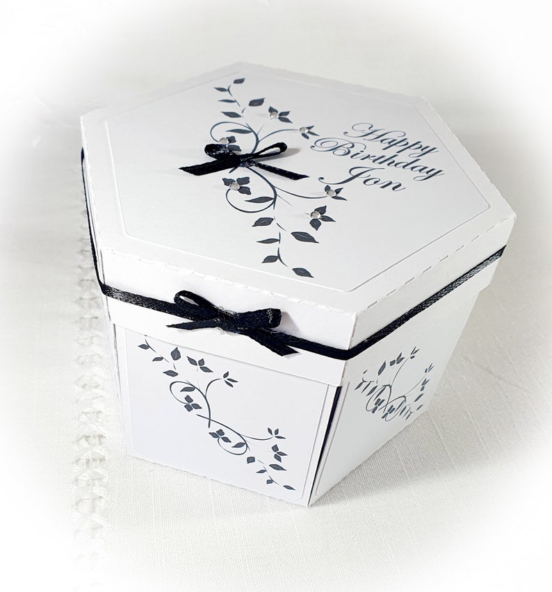 May include: White hexagonal gift box with a black ribbon and bow. The box has a floral design and the text "Happy Birthday Jon" on the top.