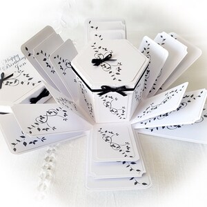 May include: A white explosion box with black floral designs and a black ribbon. The box is open and has multiple layers. The top layer has the text "Happy Birthday Jon".