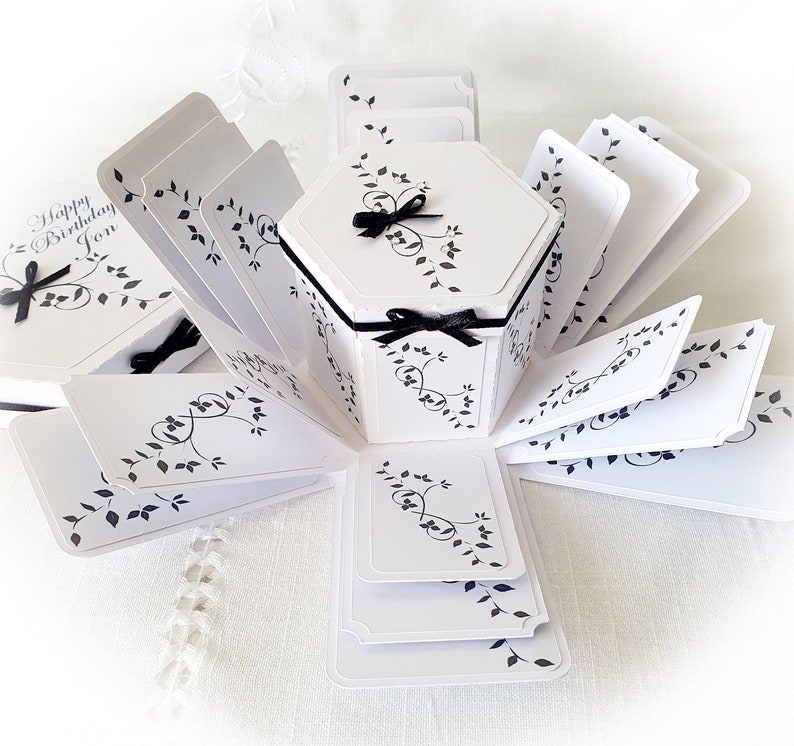 May include: A white explosion box with black floral designs and a black ribbon. The box is open and has multiple layers. The top layer has the text "Happy Birthday Jon".