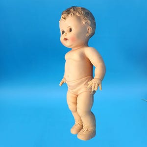 Puede incluir: Una mu&ntilde;eca beb&eacute; vintage con cara pintada, mejillas rosadas y cabello casta&ntilde;o rizado. La mu&ntilde;eca lleva un pa&ntilde;al y zapatos. La mu&ntilde;eca est&aacute; de pie sobre un fondo azul.