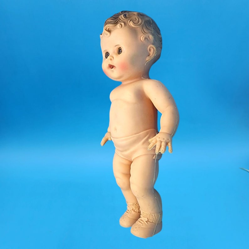 Puede incluir: Una mu&ntilde;eca beb&eacute; vintage con cara pintada, mejillas rosadas y cabello casta&ntilde;o rizado. La mu&ntilde;eca lleva un pa&ntilde;al y zapatos. La mu&ntilde;eca est&aacute; de pie sobre un fondo azul.