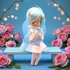 May include: A vintage angel doll with blue hair and gold accents, wearing a white dress and blue wings, sits on a light blue bench. Pink roses surround the doll against a blue backdrop with soft lighting, creating a whimsical scene.