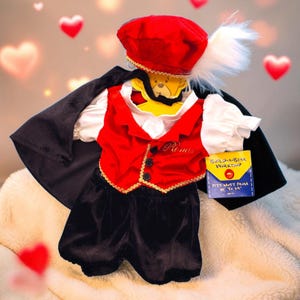 May include: A plush toy outfit featuring a red waistcoat with black buttons and the name Romeo, a black cape, black shorts, and a red beret with a white feather. A yellow tag reads Build-A-Bear Workshop and fits must from 25 to 61 cm.