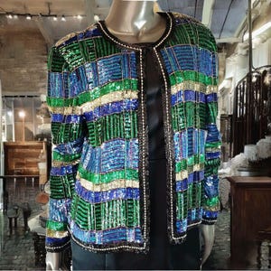 May include: A colourful sequined jacket in a patchwork design, with green, blue, and gold hues. The jacket has a round neckline and is trimmed with black and white beading. The open front design is displayed on a mannequin.