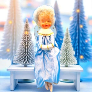 May include: A vintage angel figurine in a blue satin dress, white hair, and closed eyes, holding a small gift. The angel is set against a backdrop of white and blue Christmas trees, creating a festive holiday scene.