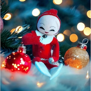 Puede incluir: Una muñeca de elfo navideño con un atuendo rojo, cuello y gorro blancos, rodeada de adornos festivos. La muñeca tiene una cara sonriente y está junto a adornos de bolas rojas y doradas, con un fondo borroso de luces.