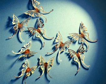 May include: Nine decorative butterfly ornaments are displayed against a blue backdrop. Each butterfly has gold-coloured filigree wings and a brown centre. The arrangement is illuminated by a spotlight.