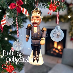 Könnte beinhalten: Ein Nussknacker-Anhänger in Form einer Figur mit Schnurrbart, bekleidet mit einer blauen Jacke mit goldenen Knöpfen, einer Krone und einer schwarzen Hose. Die Figur hängt an einer Schnur. Der Text "Holiday Delights" ist im Bild zu sehen.