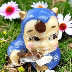 May include: A small ceramic figurine of a blue-hooded creature with brown eyes, a brown spot on its forehead, and a brown stick in its hand.